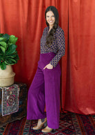 woman with long hair wears purple corduroy wide leg pants with her hand in pockets, paired with a long sleeve cotton blouse 