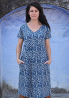 Woman poses in blue based floral print cotton dress with short sleeves and a V-neckline, with her hands in pockets