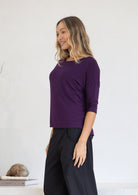 Woman wearing a purple top with 3/4 sleeves and dark pants against a white background