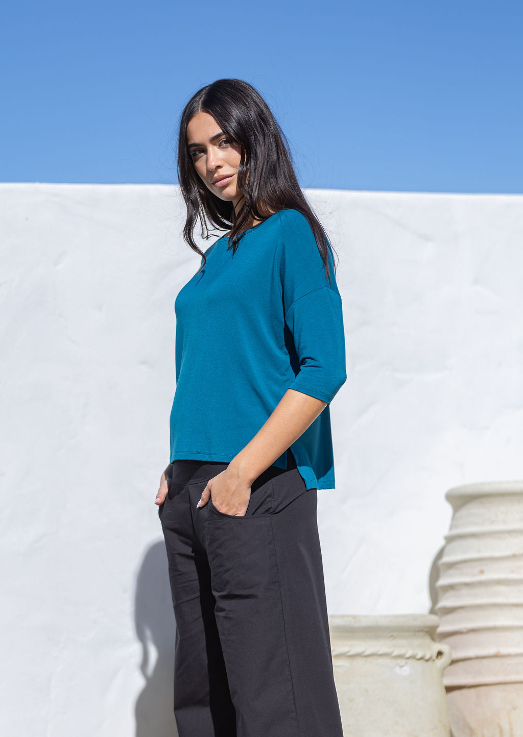 Woman wearing a teal top and black pants against a white wall with blue sky.
