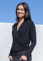 Woman wearing a black long sleeve crossover top against a blue sky and white wall.