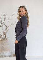 Woman stands side on in a dark grey long sleeve crossover top with hand in pocket of pants