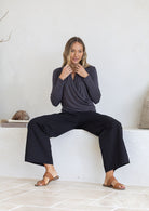 Woman sitting on a white ledge wearing a dark grey crossover long sleeve top and black pants.