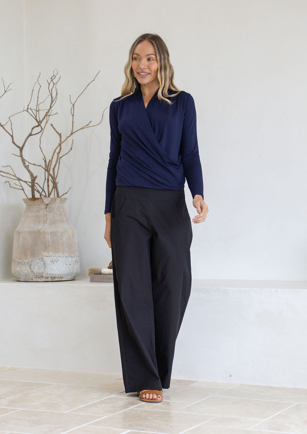 Woman wearing a navy blue long sleeve false wrap top and black pants standing in a minimalistic room