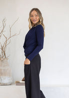 Woman stands side on in navy blue long sleeve cross over top with hand in pocket of pants