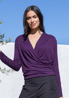 Woman wearing a purple long sleeve false wrap top against a blue sky and white wall.
