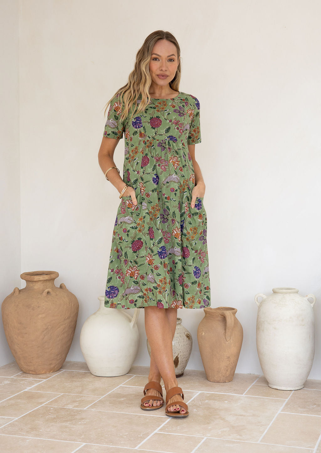 Woman wearing a green based floral print cotton dress with hands in pockets.