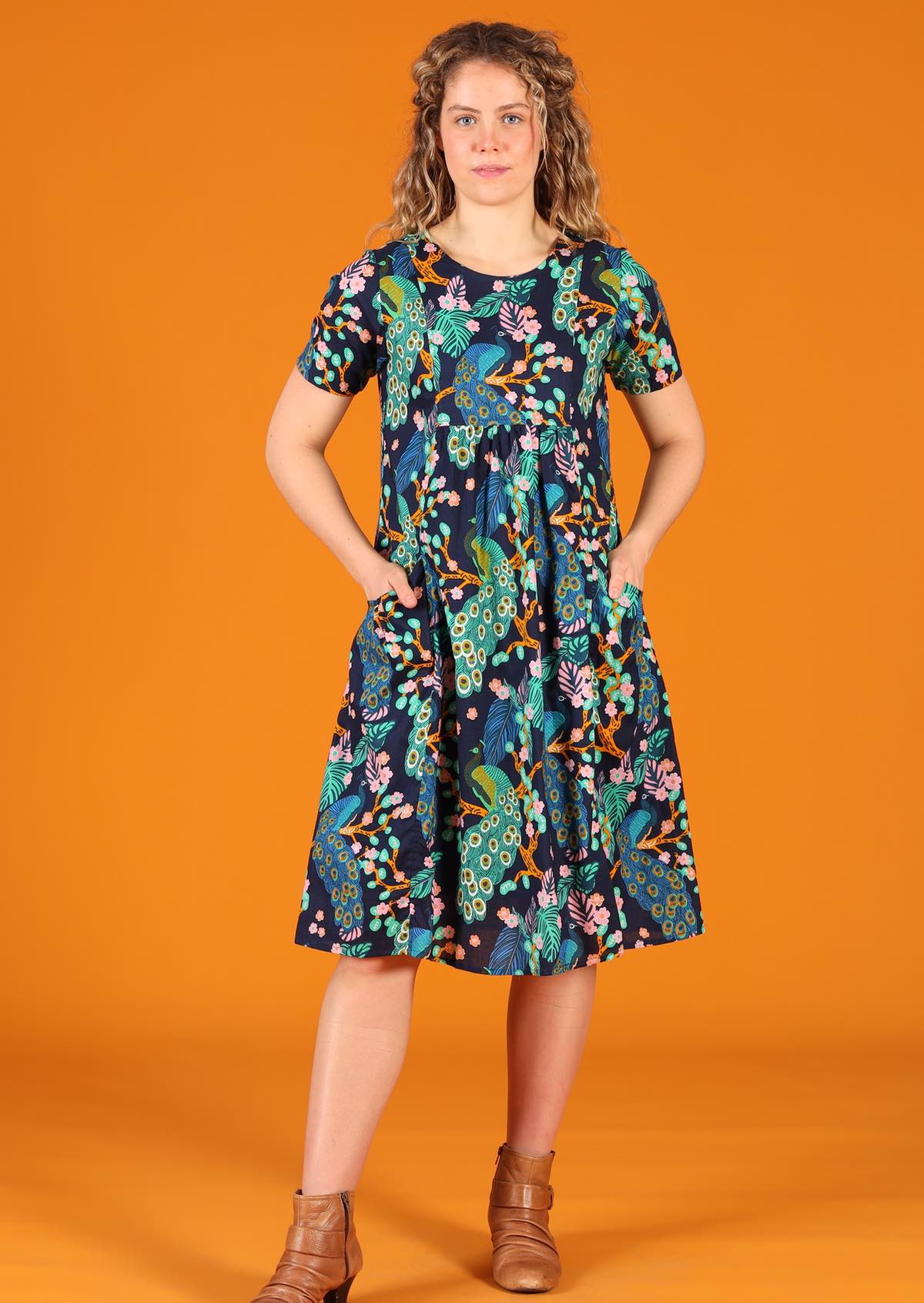Model wearing blue green peacock print cotton dress with hands in pockets 