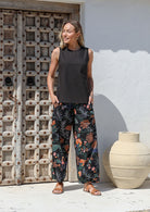 Woman standing in front of wooden door with both the hands in pockets wears 100% cotton wide leg black pants in queen protea print