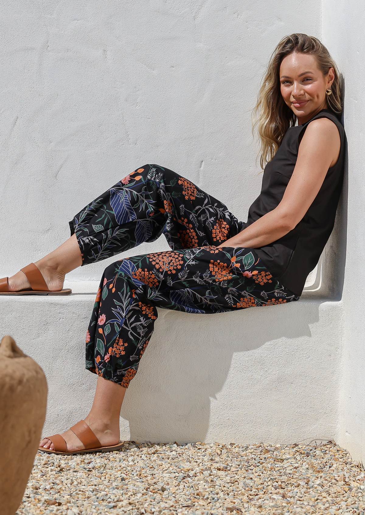 Woman sitting on a white concrete bench wall wearing black botanic print wide leg cotton pants and a black sleeveless top.