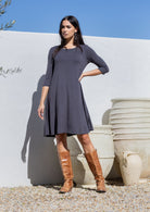 Woman wearing a dark gray stretch jersey dress and brown knee-high boots standing against a white wall with large pots.