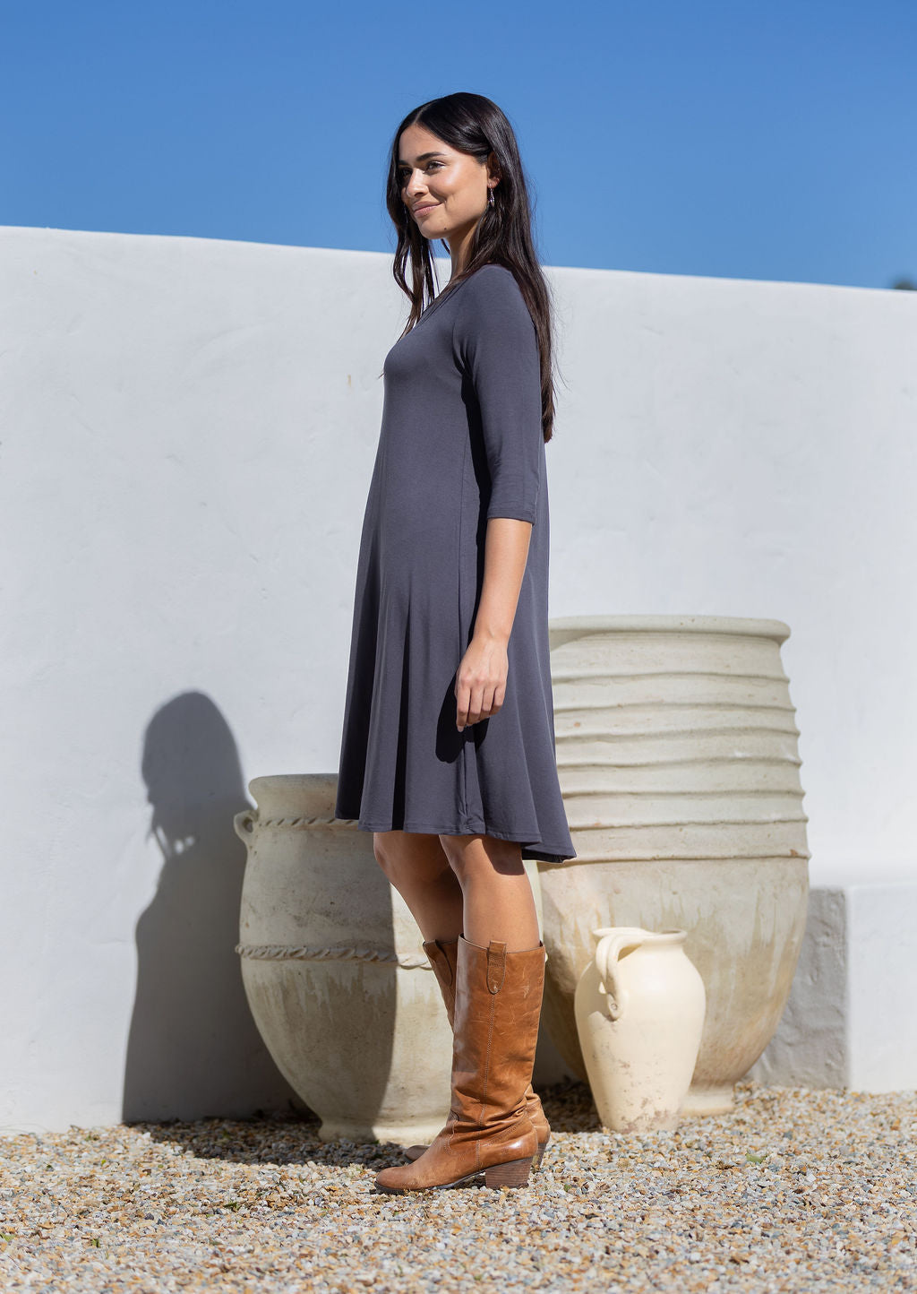 Woman in a grey jersey dress and brown boots standing in front of white pots against a blue sky.