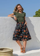 Woman stands with one hand in pocket wearing a basic green top and black botanic print, button through cotton skirt with box pleats and belt loops 
