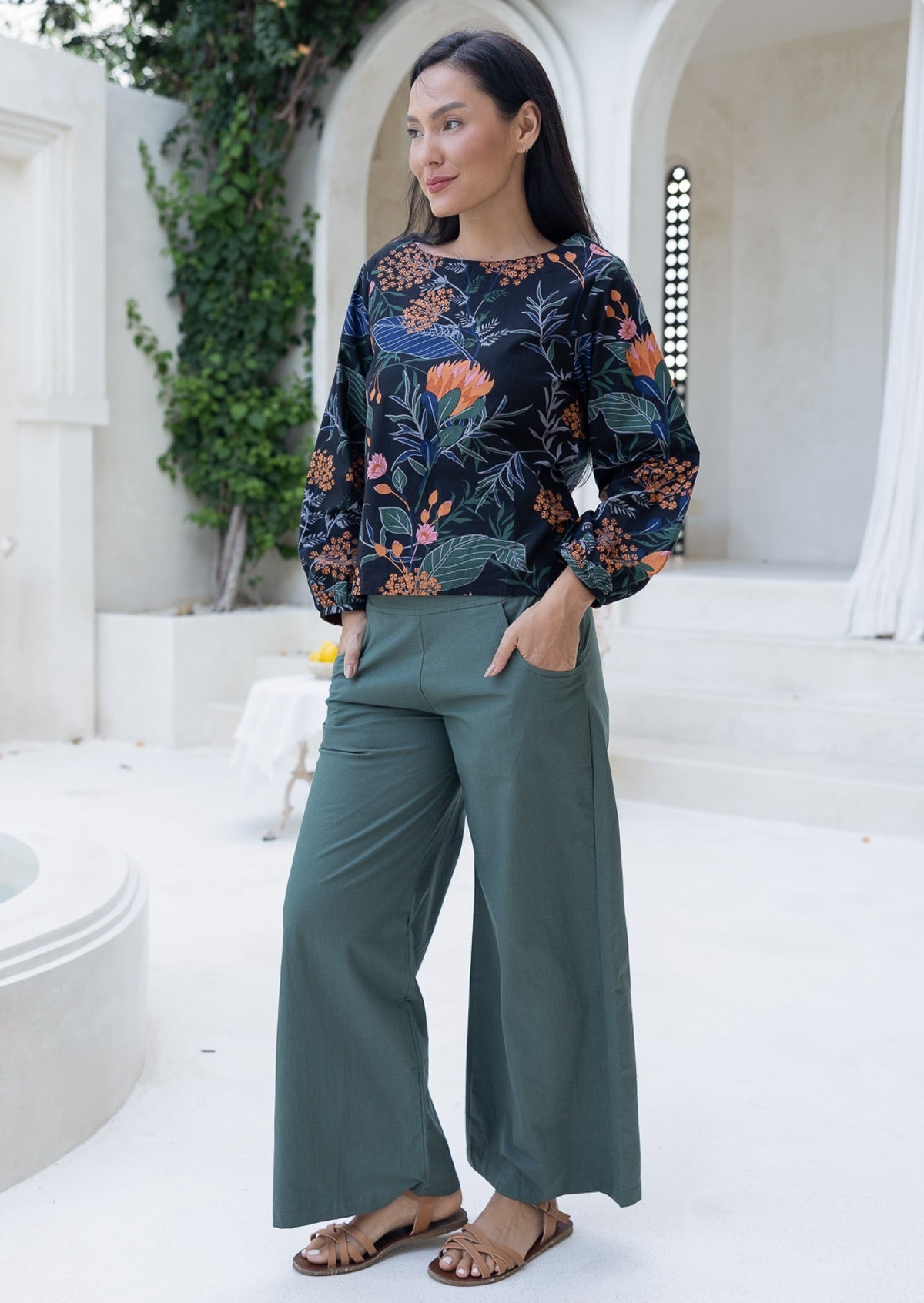 Woman wearing a loose fitting botanical printed cotton top and green pants standing in a white architectural setting