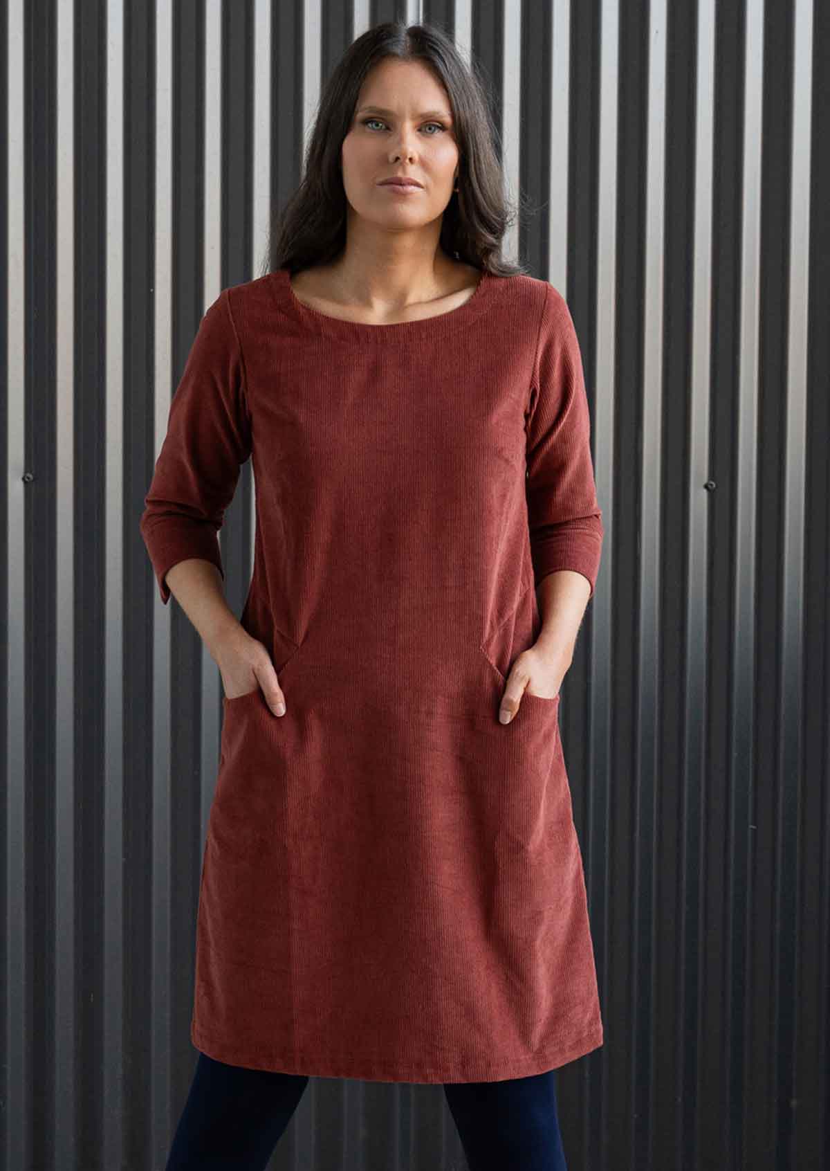 Woman wearing rust coloured corduroy tunic with sleeves over navy blue leggings and boots with hands in pockets 