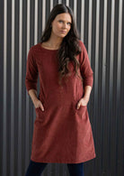 Woman wearing rust coloured cotton corduroy dress with sleeves over navy blue leggings and boots with hands in pockets 