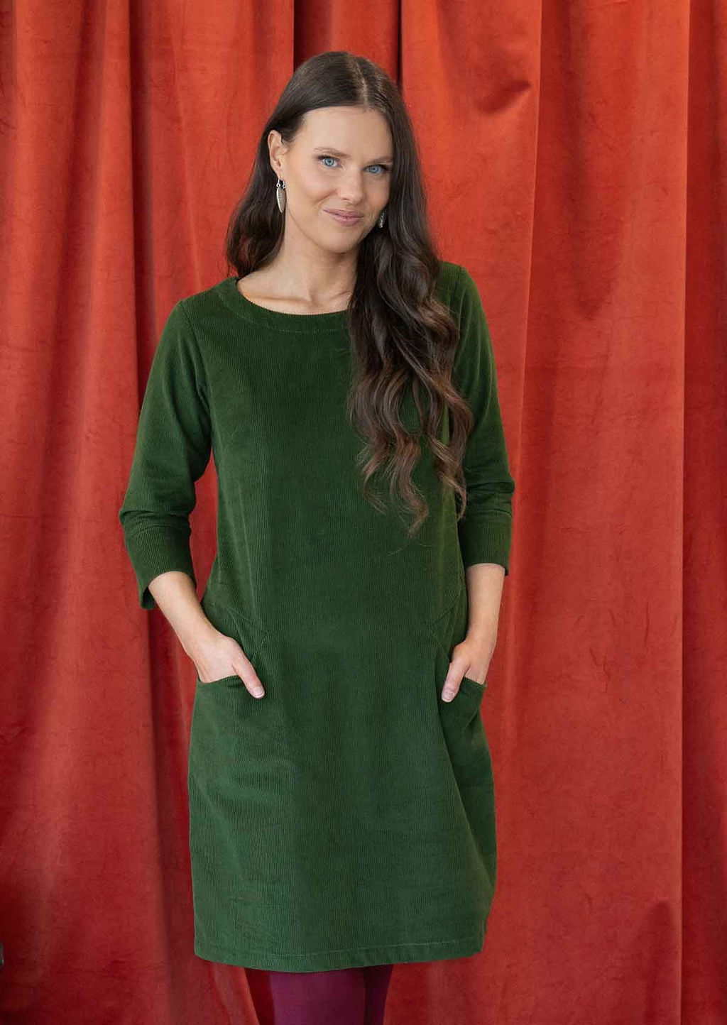 Woman wearing green corduroy A-line tunic dress with 3/4 sleeves with hands in pockets 