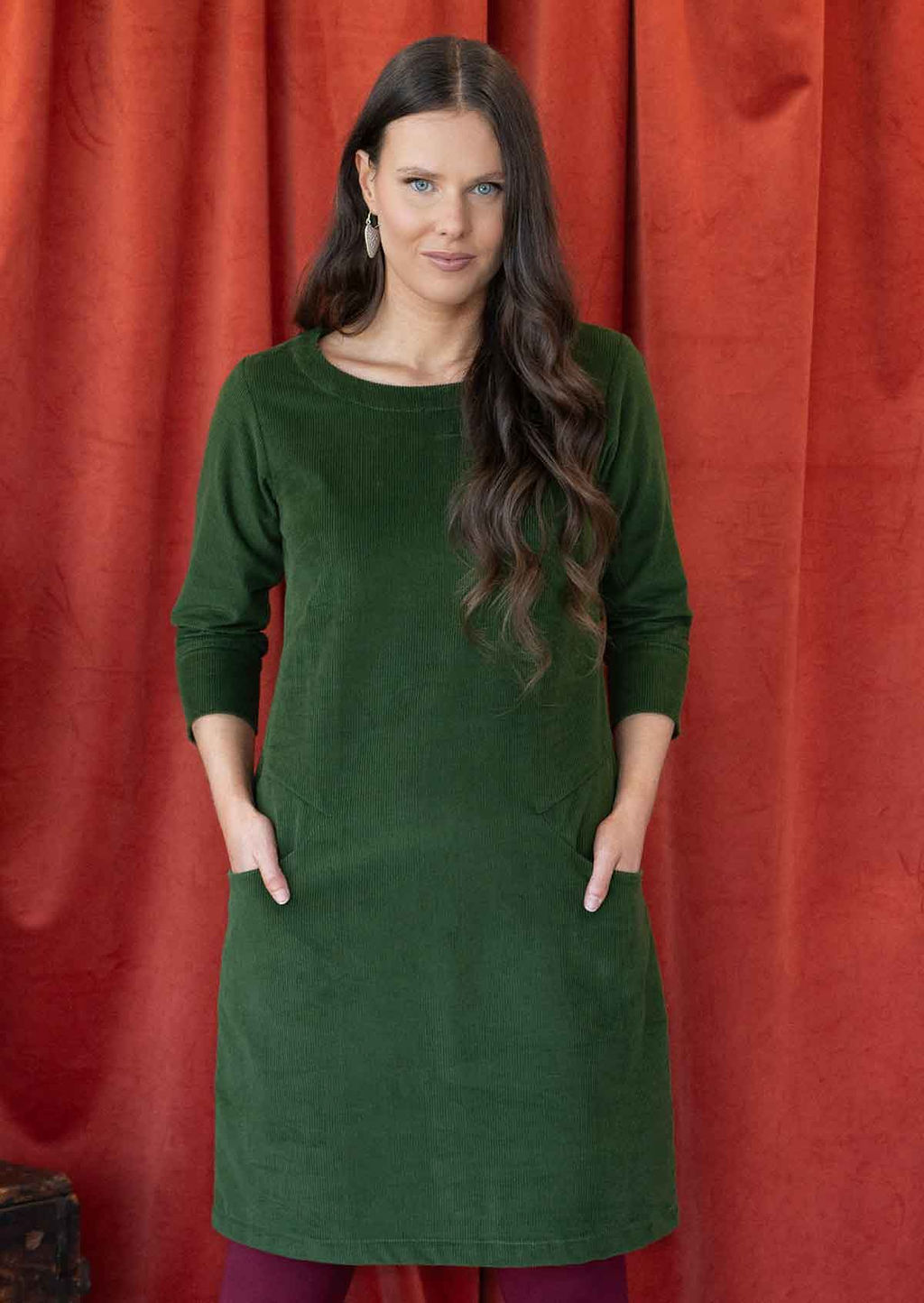 Woman wearing green cotton corduroy A-line dress over leggings with 3/4 sleeves with hands in pockets 