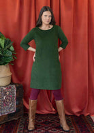 Woman wearing green corduroy A-line dress with 3/4 sleeves over maroon leggings and tan boots