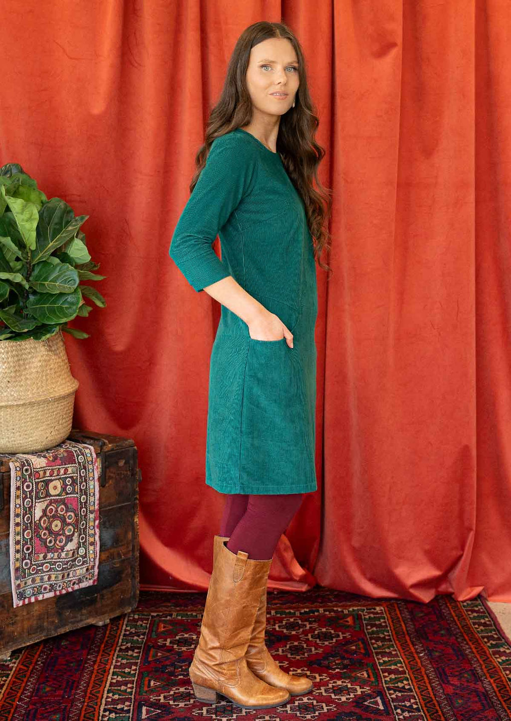 Woman wearing teal corduroy dress over maroon leggings and long boots hand in pocket