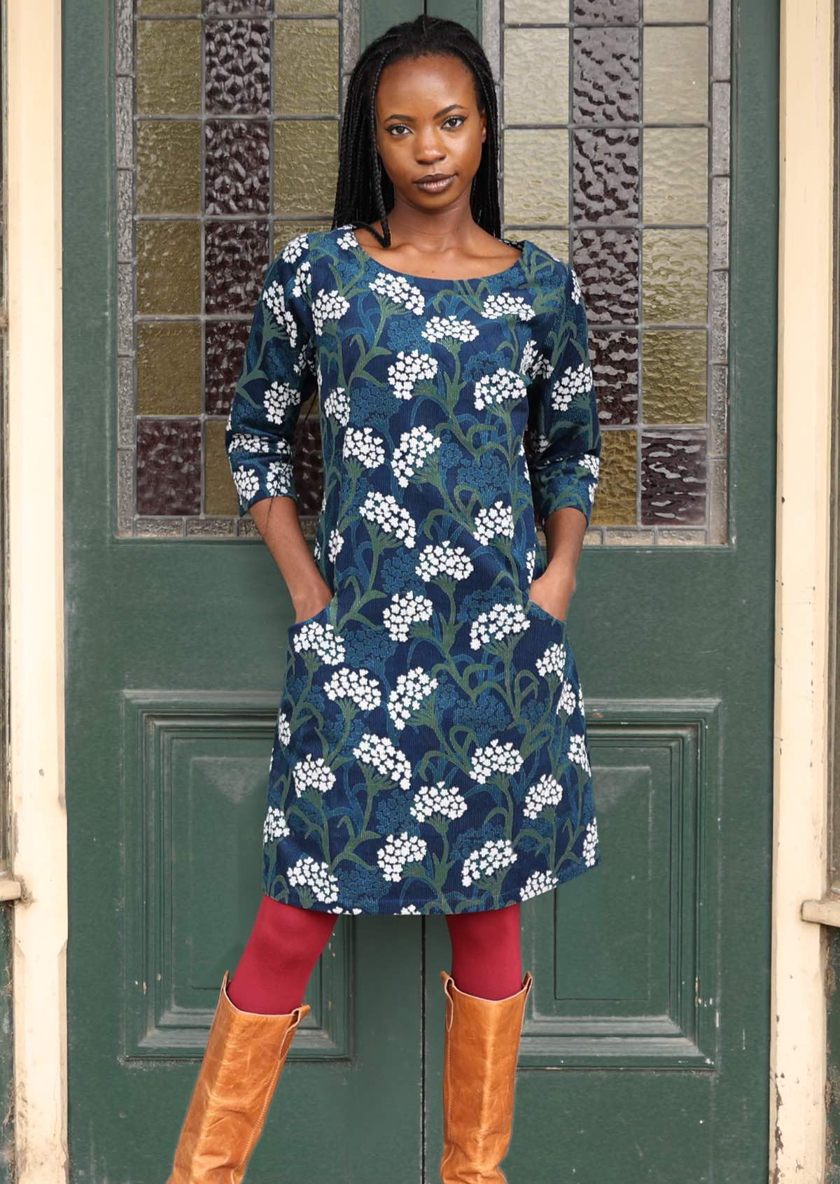 Woman wears wide neck 3/4 sleeve corduroy dress with hands in pockets, paired with maroon leggings and boots