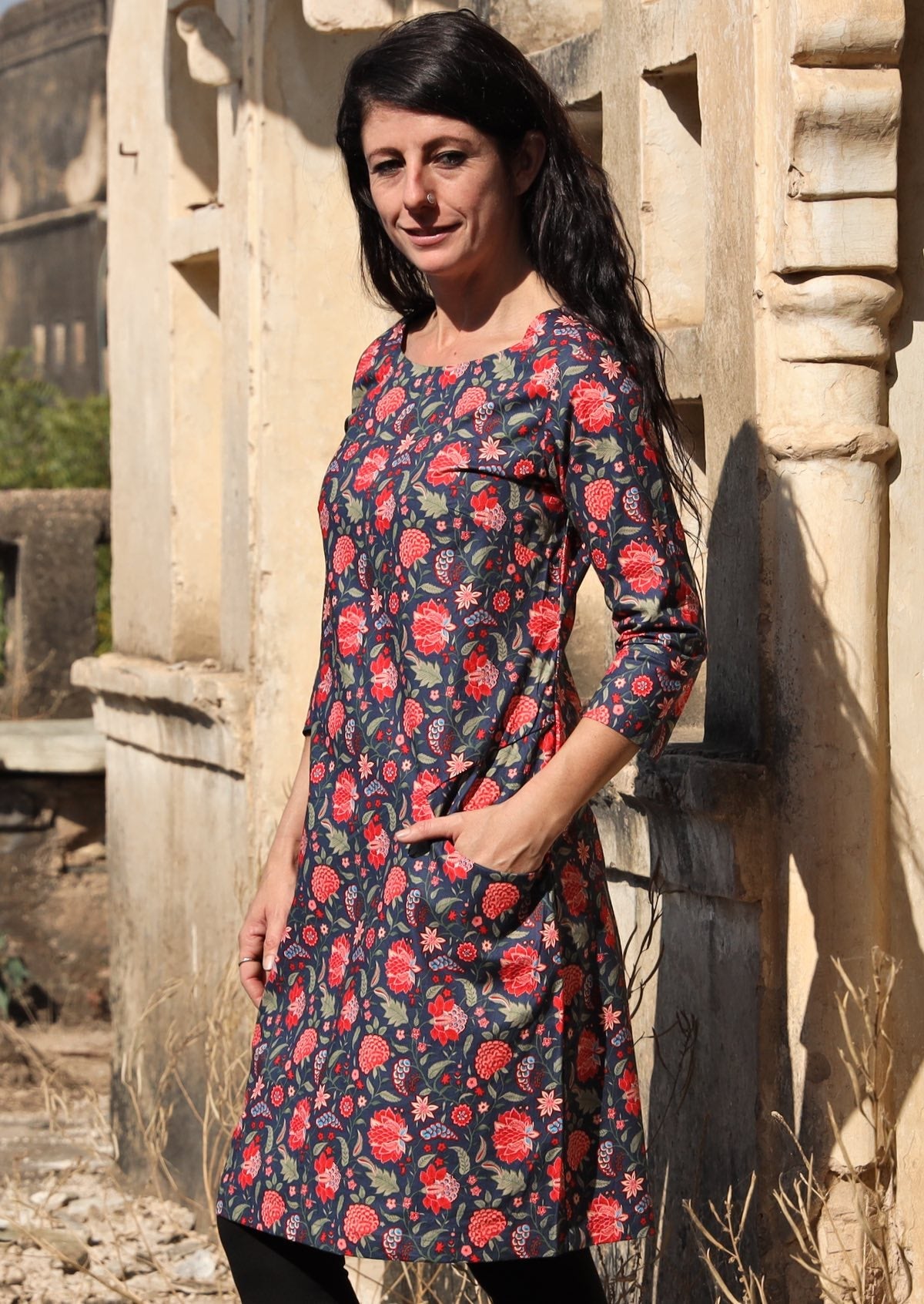 Woman standing to the side with hand in pocket wearing long sleeve 100 percent cotton dress with deep blue base and raspberry red floral print 
