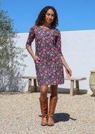 Woman wearing a navy and red floral dress with brown boots standing outdoors against a white wall.