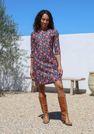 Woman wearing a navy cotton floral dress and brown boots standing outdoors with a white wall and tree in the background.