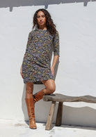 Woman wearing a black cotton patterned dress and brown knee-high boots leaning on rustic stool 