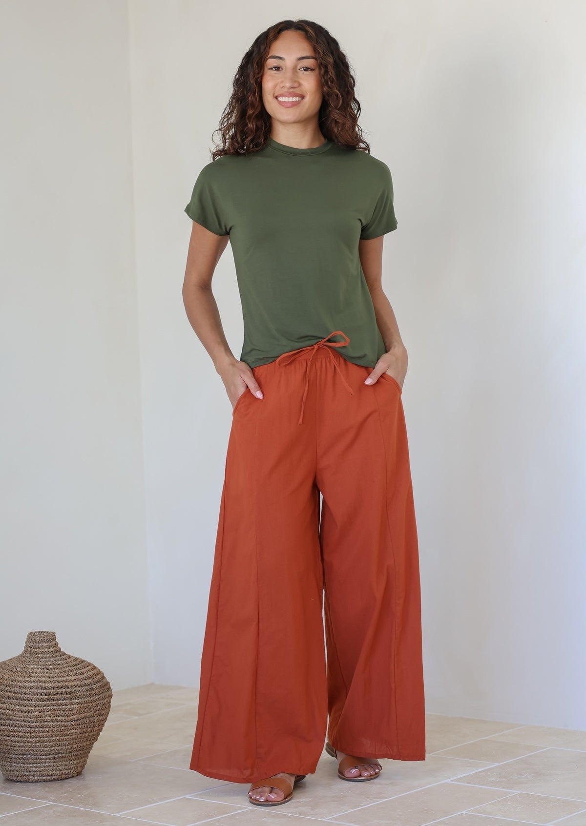 Woman wearing a green t-shirt and rust colored wide-leg cotton pants with drawstring tie and hands in pocket standing against the white wall.