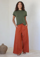 Woman wearing a green t-shirt and rust colored wide-leg cotton pants with drawstring tie and hands in pocket standing against the white wall.