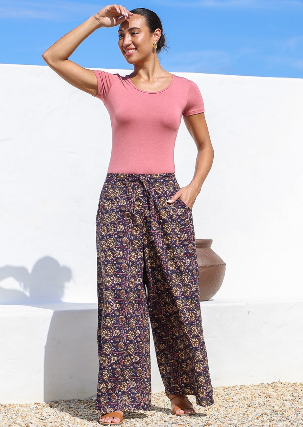 Model stands in sun wearing high waisted, relaxed fit cotton pants with drawstring and a hand in pocket. 