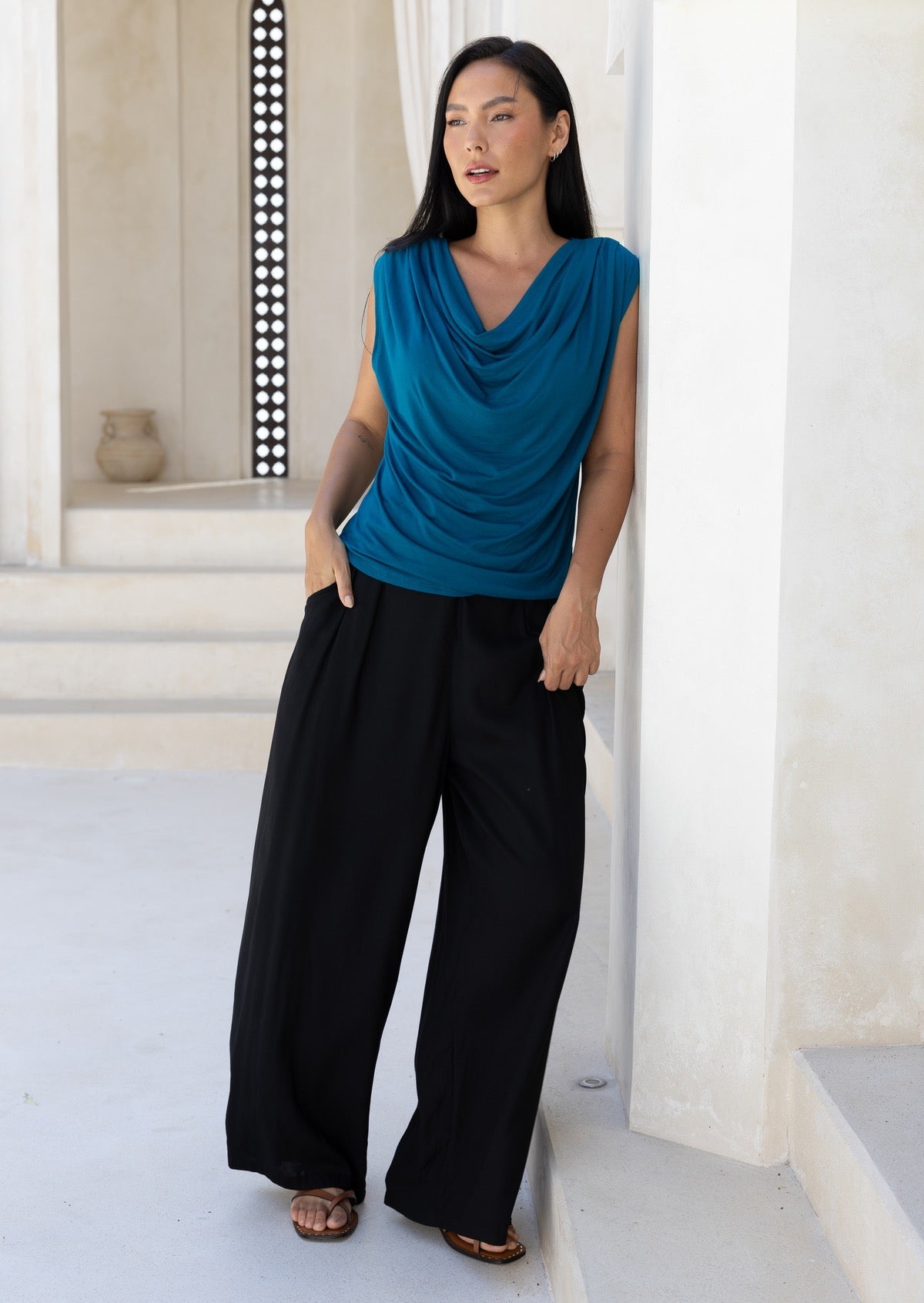 Woman wearing a blue sleeveless top and black pants standing in a bright, minimalistic room.