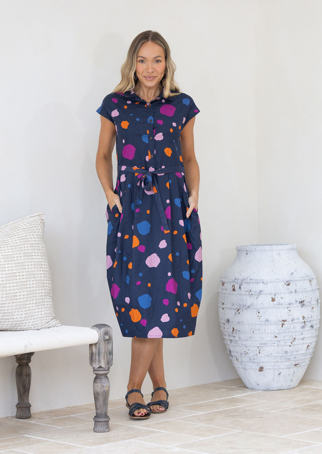 Woman wearing a navy based cotton day dress with colourful polka dots in a minimal indoor setting, with hands in pockets