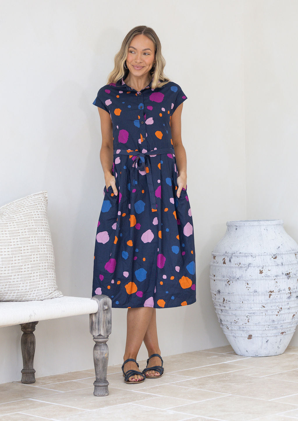 Woman wearing a blue based colourful spotty cotton dress with buttoned bodice and collar, with her hands in pockets