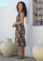 Model stands sideways near ceramic pots, wearing a midlength, shirt style floral print cotton dress with a black base and yellow, blue, and grey florals