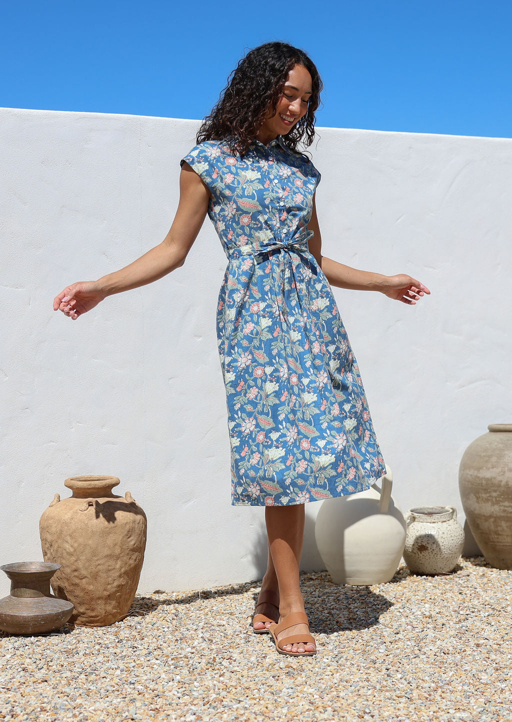 Model poses in a retro style cotton dress featuring a dusty blue based sweet floral print, buttoned bodice, fabric waist tie and cap sleeves