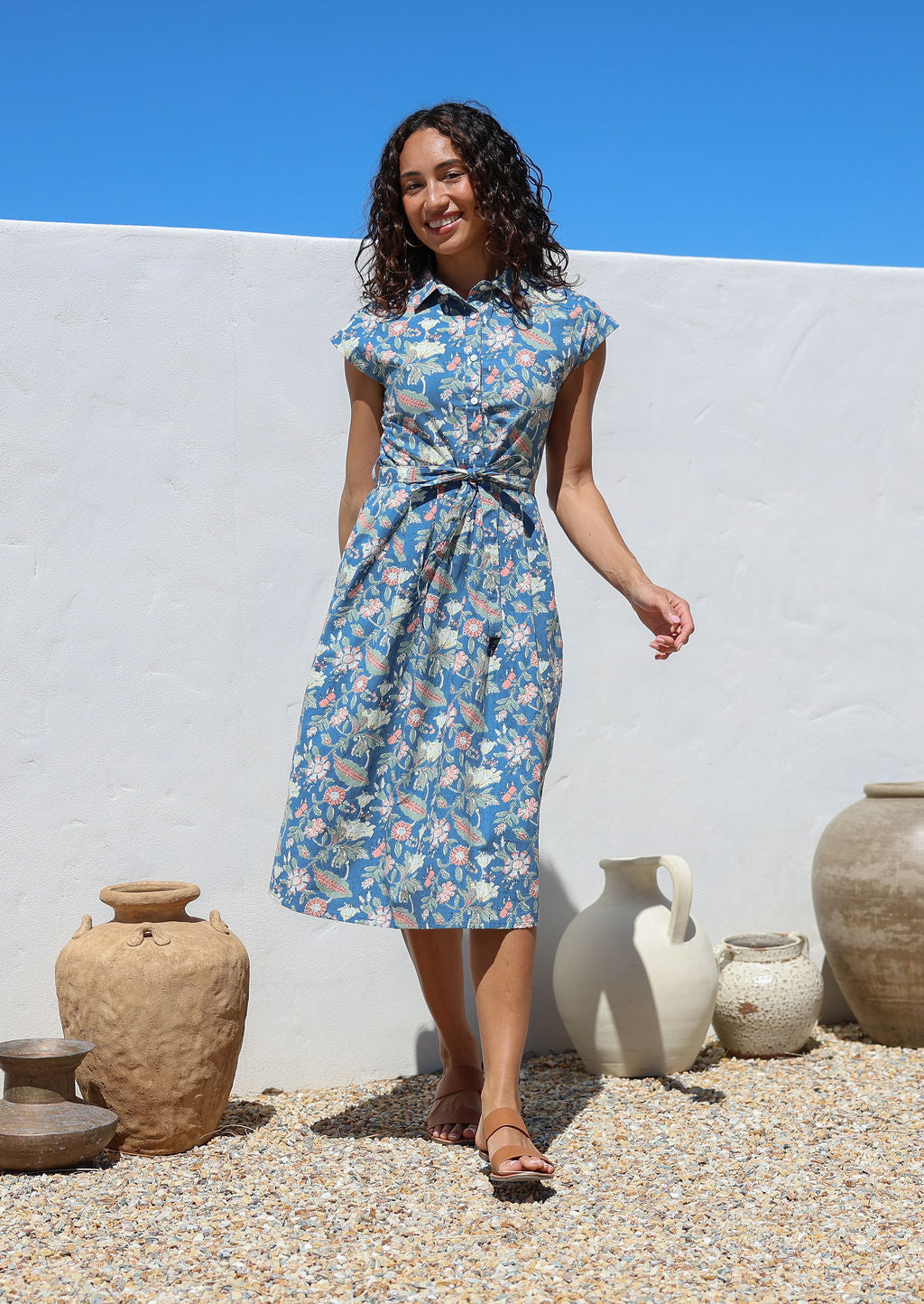 Woman in a blue floral shirt style cotton dress with buttoned bodice standing outdoors with a white wall and decorative pots in the background