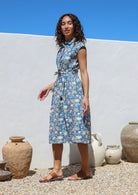 Model stands sideways wearing a floral printed blue shirt style cotton dress with collar, buttoned bodice, pockets and cap sleeves