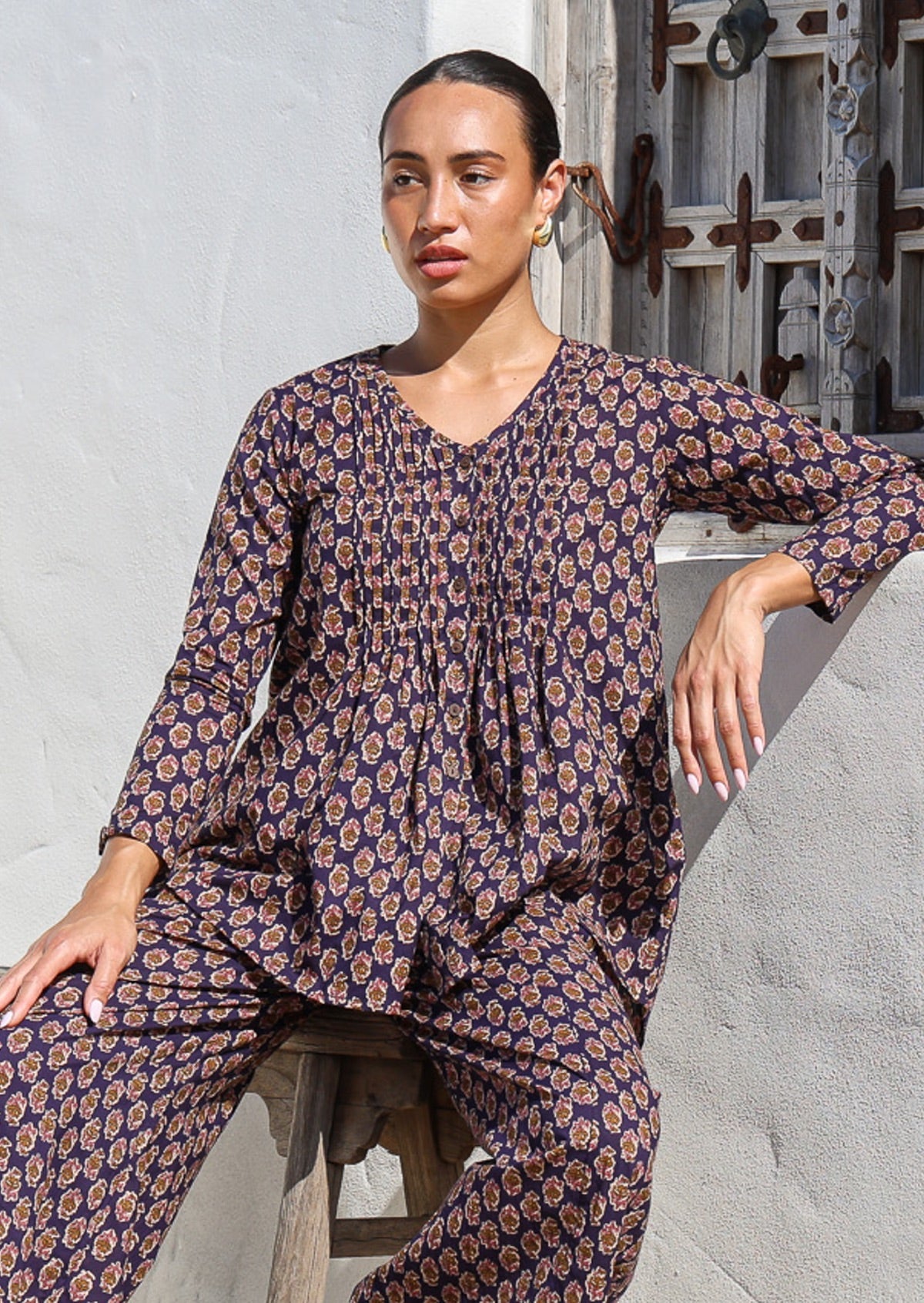 Model sitting on a wooden stool wears lightweight 100% cotton long sleeve, button through top in dark purple based floral print.