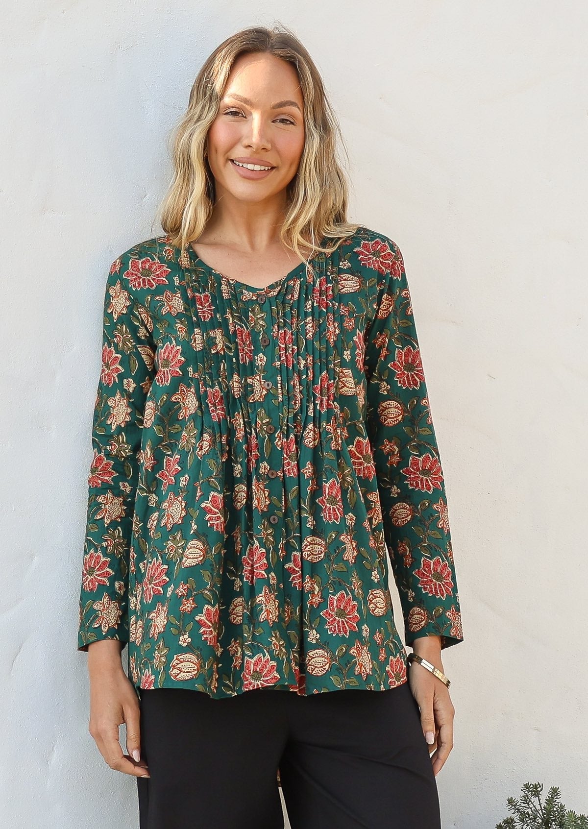 Woman wearing a green based cotton floral long sleeve relaxed fit blouse with a V-neckline, against a white wall