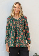 Woman wearing a green based cotton floral long sleeve relaxed fit blouse with a V-neckline, against a white wall
