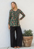 Woman wearing a cotton floral long-sleeve button through shirt and black pants standing next to a potted plant.
