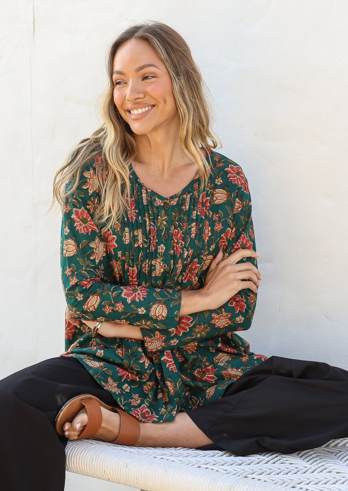 Woman wearing a cotton floral long-sleeve shirt with a V-neckline, sitting against a white wall.