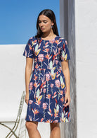 Woman wears loose fit 100% cotton dress with round neckline and pockets in native pink and green floral print on navy base.