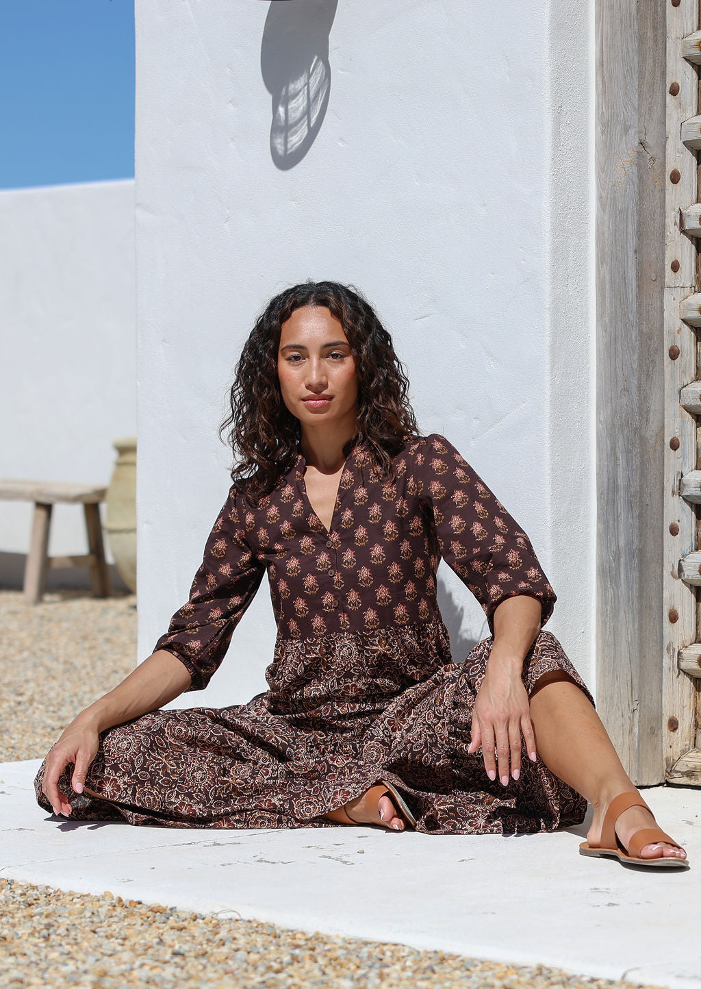Woman in a Indian cotton maxi dress sitting on the ground with a white wall and blue sky in the background.