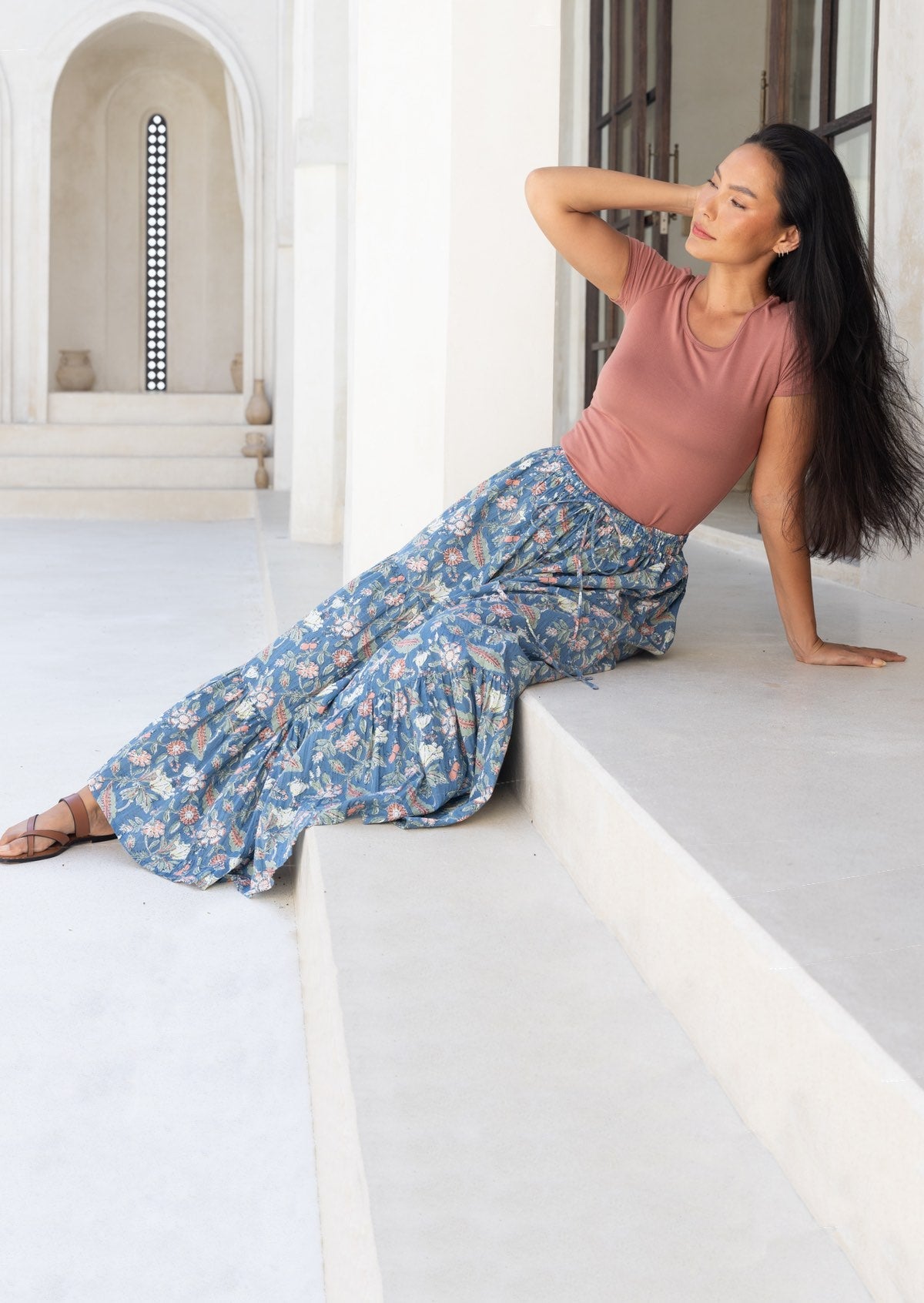 Woman sitting on a ledge wearing a pink top and blue cotton floral maxi skirt in a bright, architectural setting.