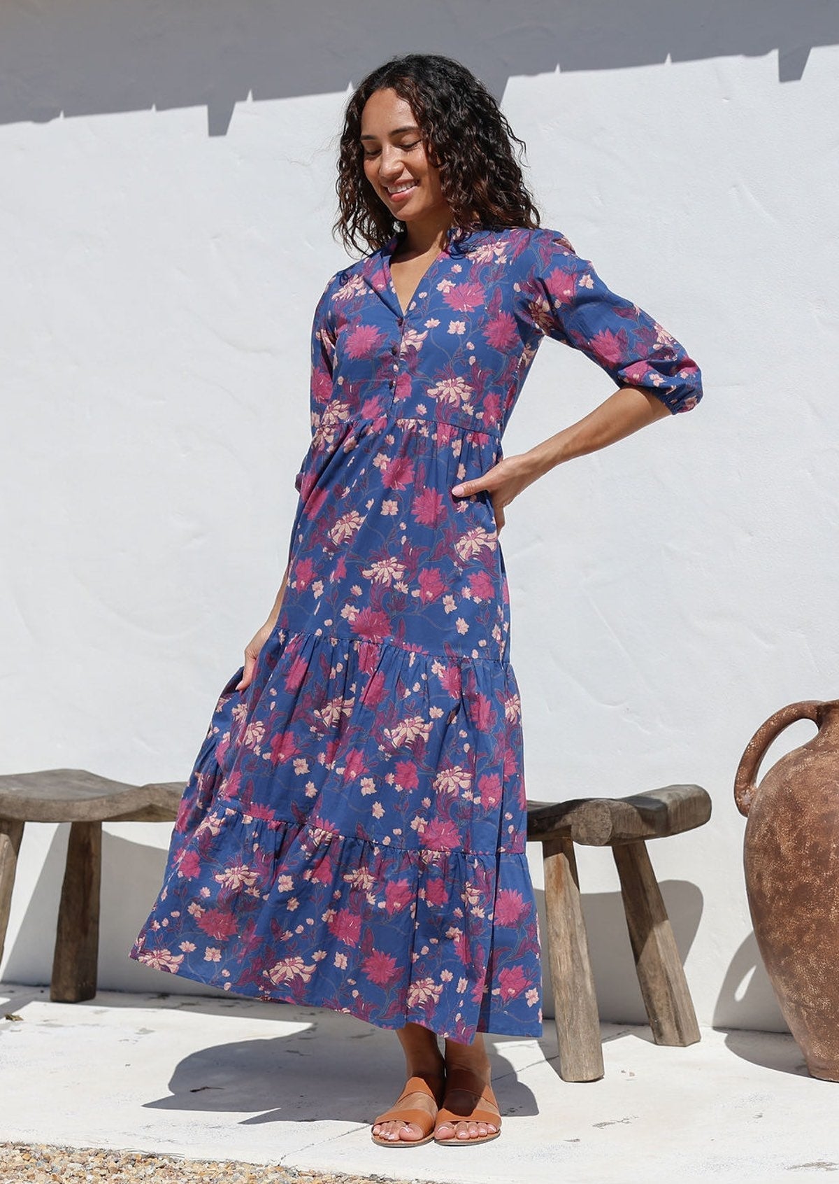 Woman wearing a navy blue floral maxi dress standing against a white wall with a wooden bench and vase