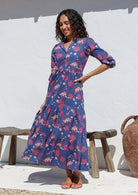 Woman wearing a navy blue floral maxi dress standing against a white wall with a wooden bench and vase