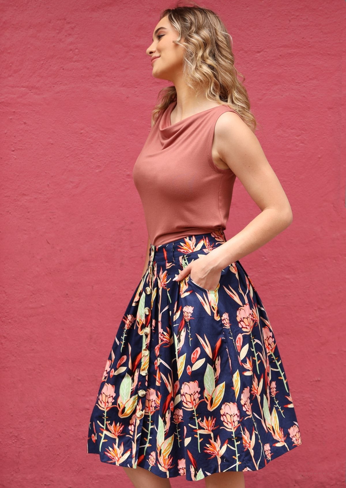 Woman wearing a pink top and floral cotton skirt with pink, orange and green floral print on a blue base, against a pink wall